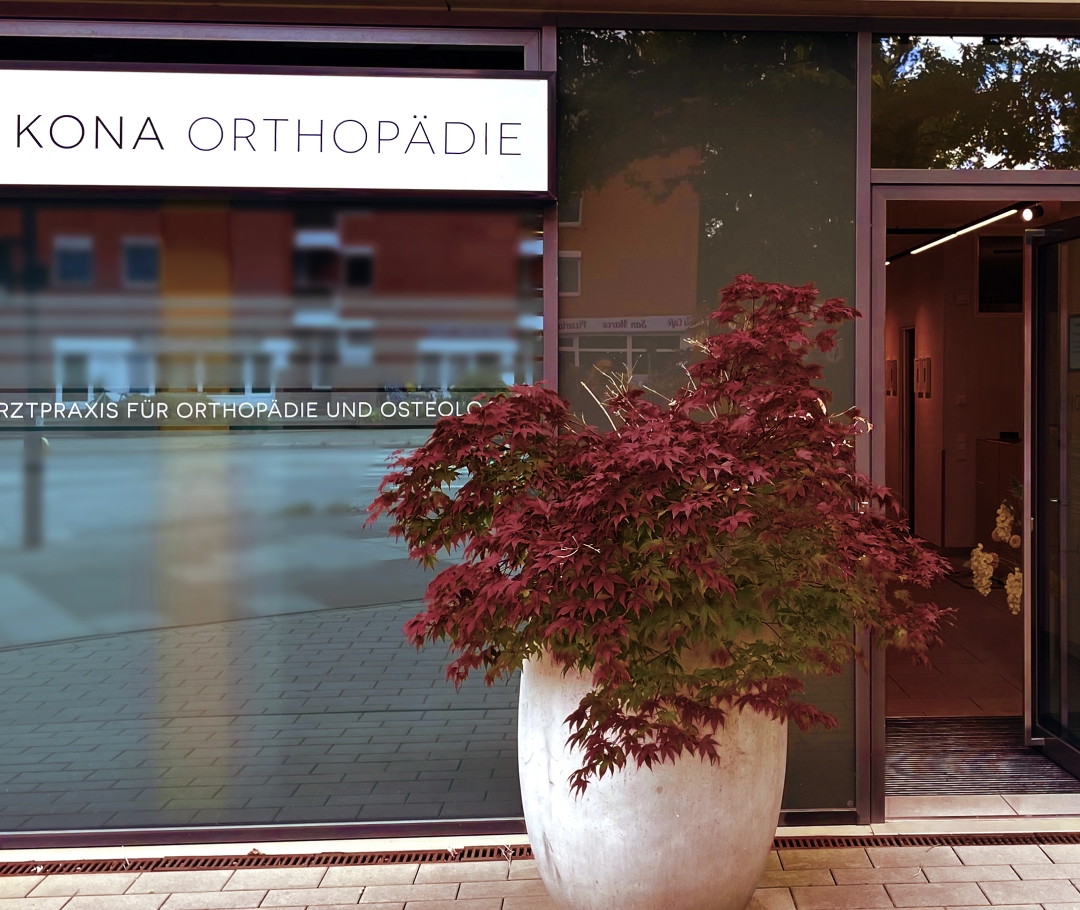 Eingang der Praxis „KONA Orthopädie“ mit rotem Ahorn im großen Topf vor der Tür.
